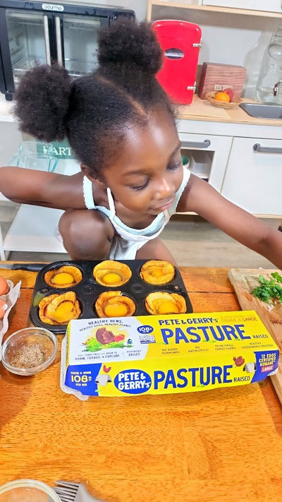 A little girl with a muffin tray in front of her. There's a carton of Pete & Gerry's eggs on the table