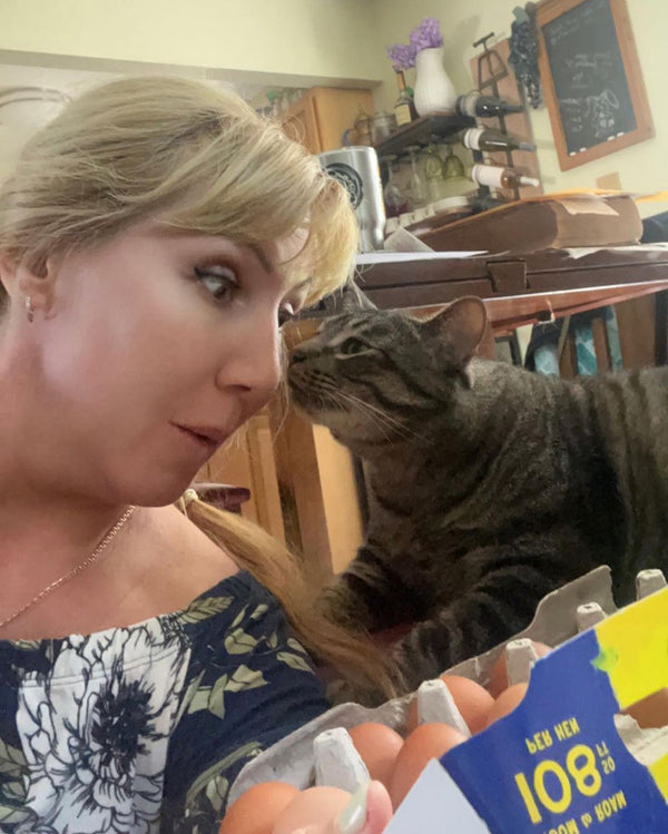 A woman getting a kiss from a gray cat. She's holding a carton of Pete & Gerry's eggs