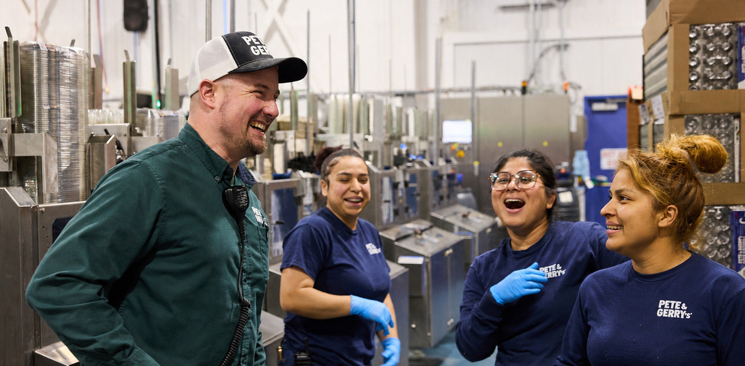 Group of people in a factory setting, smiling and interacting with each other.